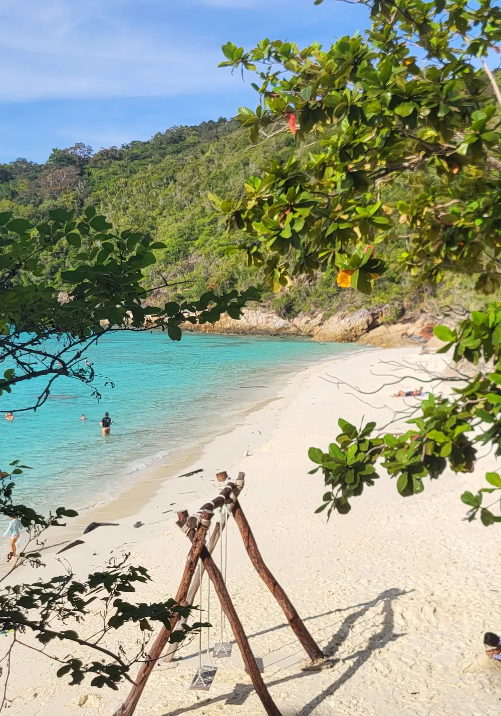 A white-sand beach on Perhentian Kecil with turquoise water, a wooden swing frame and a forested headland under a blue sky