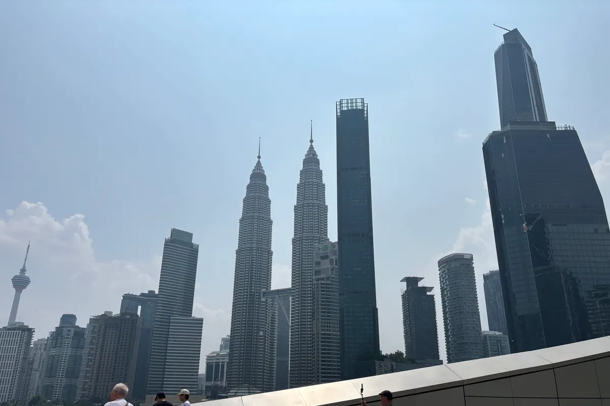 The Kuala Lumpur skyline under a hazy sky, with the Petronas Twin Towers, Merdeka 118 and KL Tower seen from a rooftop park