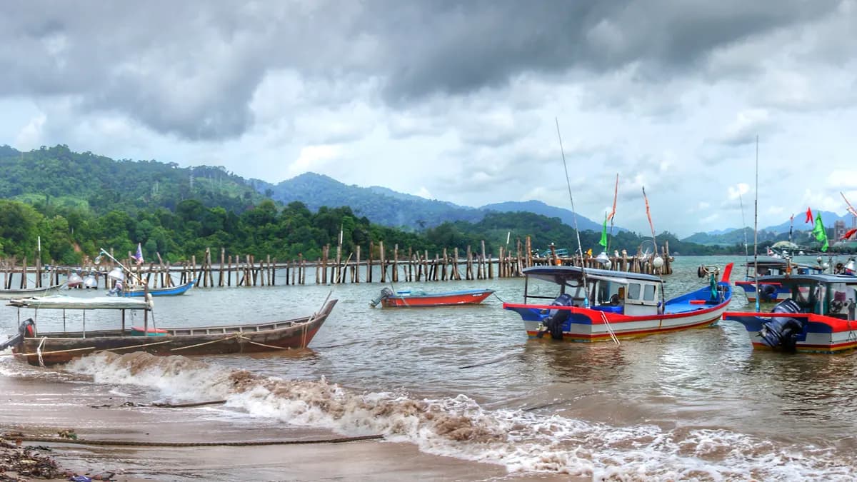 Fishing boats moored beside a wooden pier at Black Sand Beach on Langkawi, with forested hills and heavy cloud behind