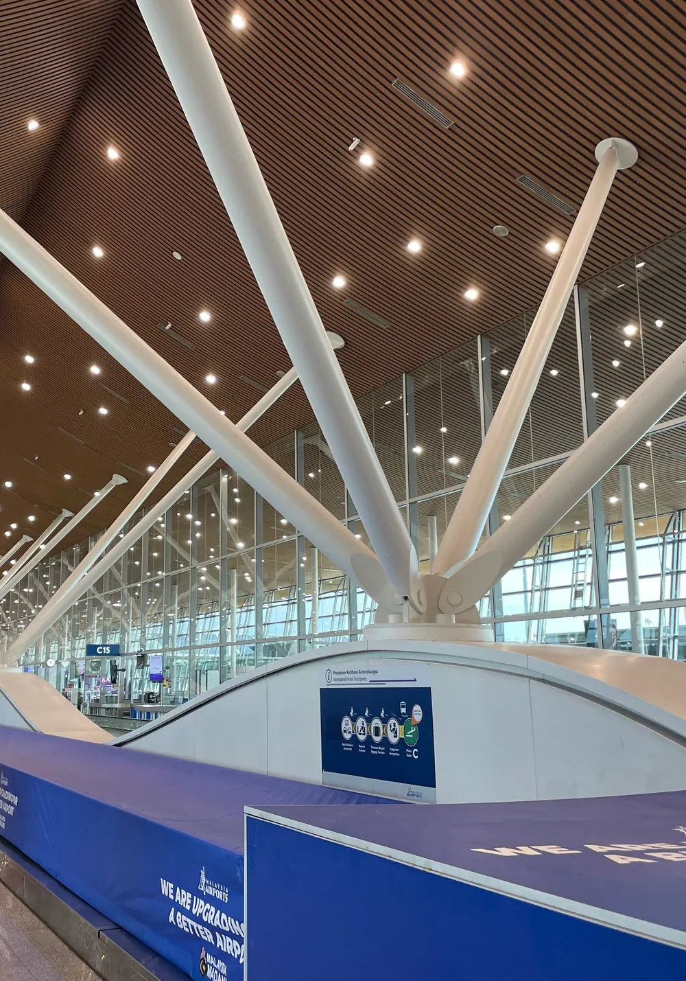 White steel columns branching under the timber ceiling of a concourse at Kuala Lumpur International Airport, with travellers walking beside the glass wall
