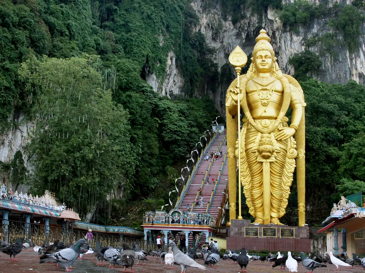 The golden statue of Lord Murugan beside the long, colourful staircase climbing the limestone cliff to the temple cave at Batu Caves, with pigeons on the forecourt