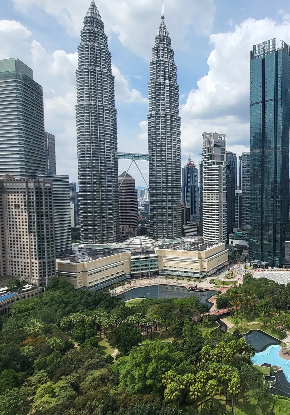 The Petronas Twin Towers and the surrounding skyscrapers of Kuala Lumpur rising above the trees, lake and paths of KLCC Park under a cloudy sky