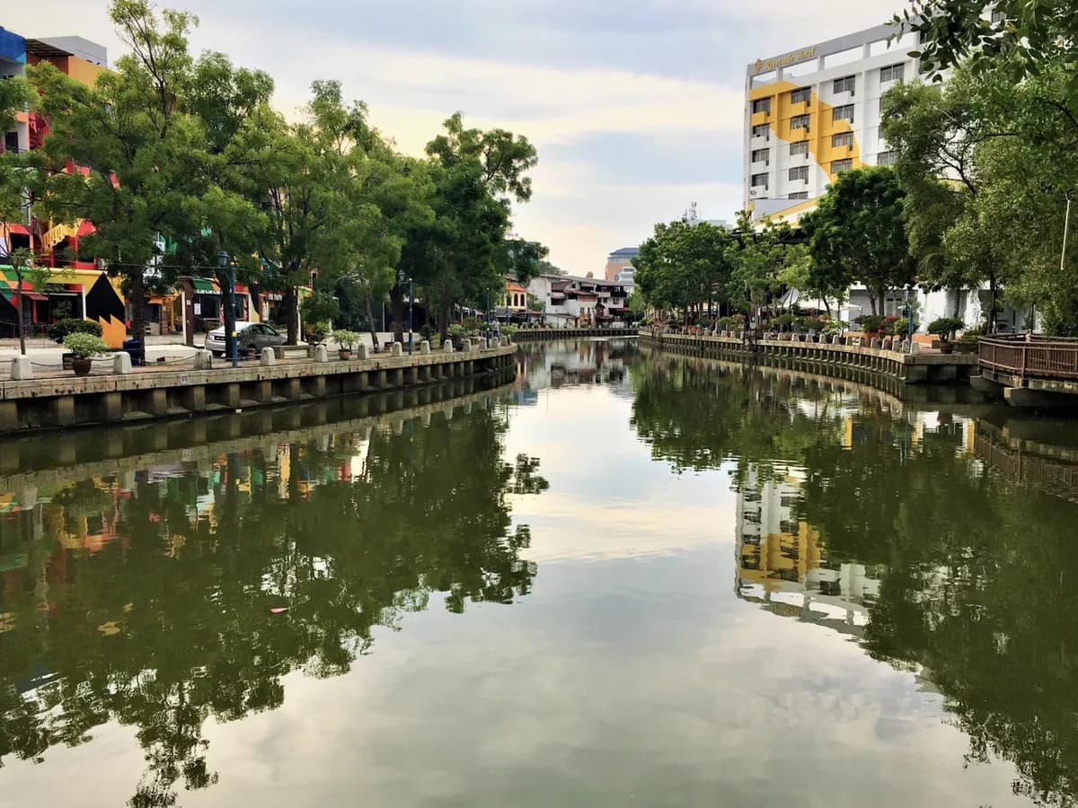 The Melaka River between its stone embankments, with trees, painted riverside shophouses and a hotel reflected in the calm water