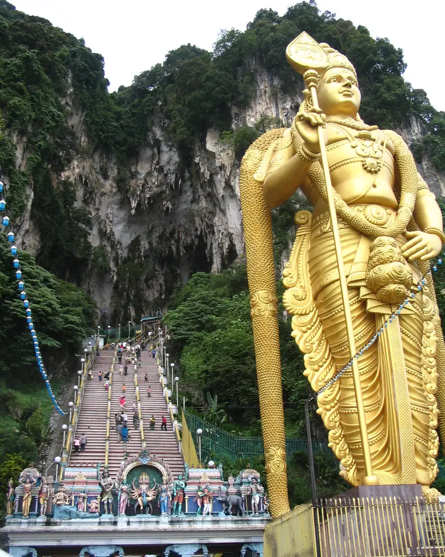 The tall golden Murugan statue beside the long colorful staircase that climbs to the limestone cliffs and temple cave at Batu Caves