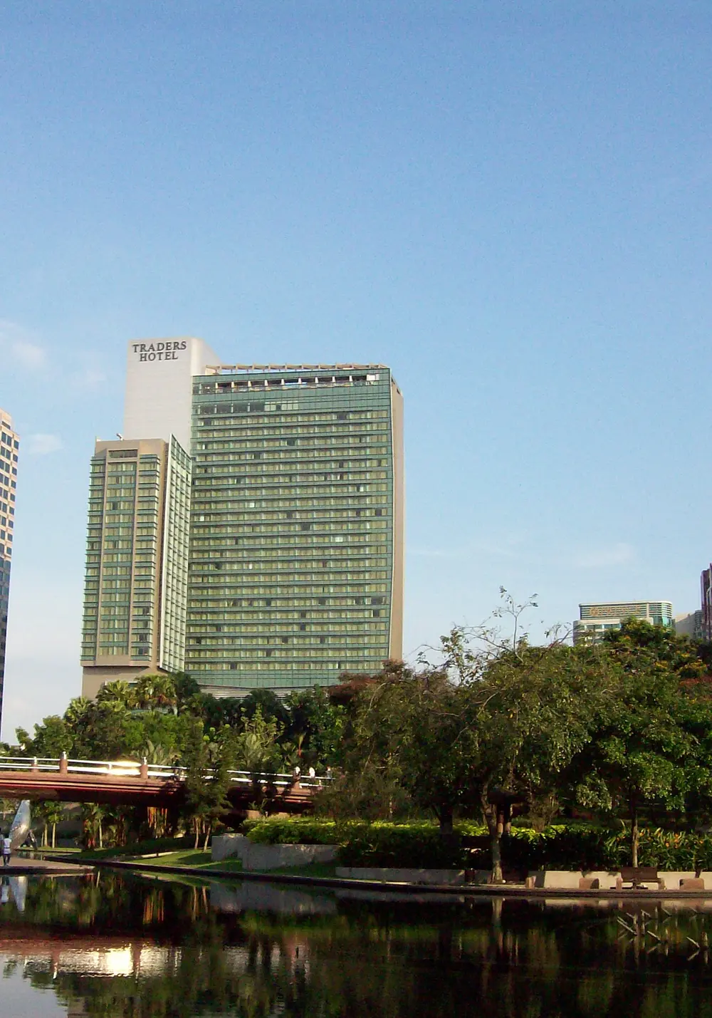 Tall hotel and office towers rise above trees beside a still lake in KLCC Park, Kuala Lumpur, under a blue sky