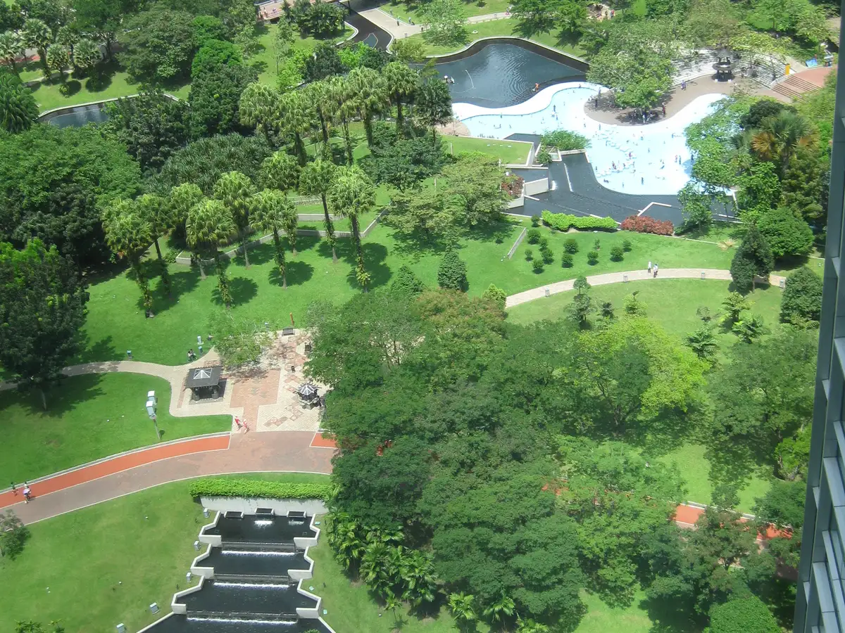 View over KLCC Park in Kuala Lumpur from above, with curving walking paths, green lawns, tall trees and a curved pool