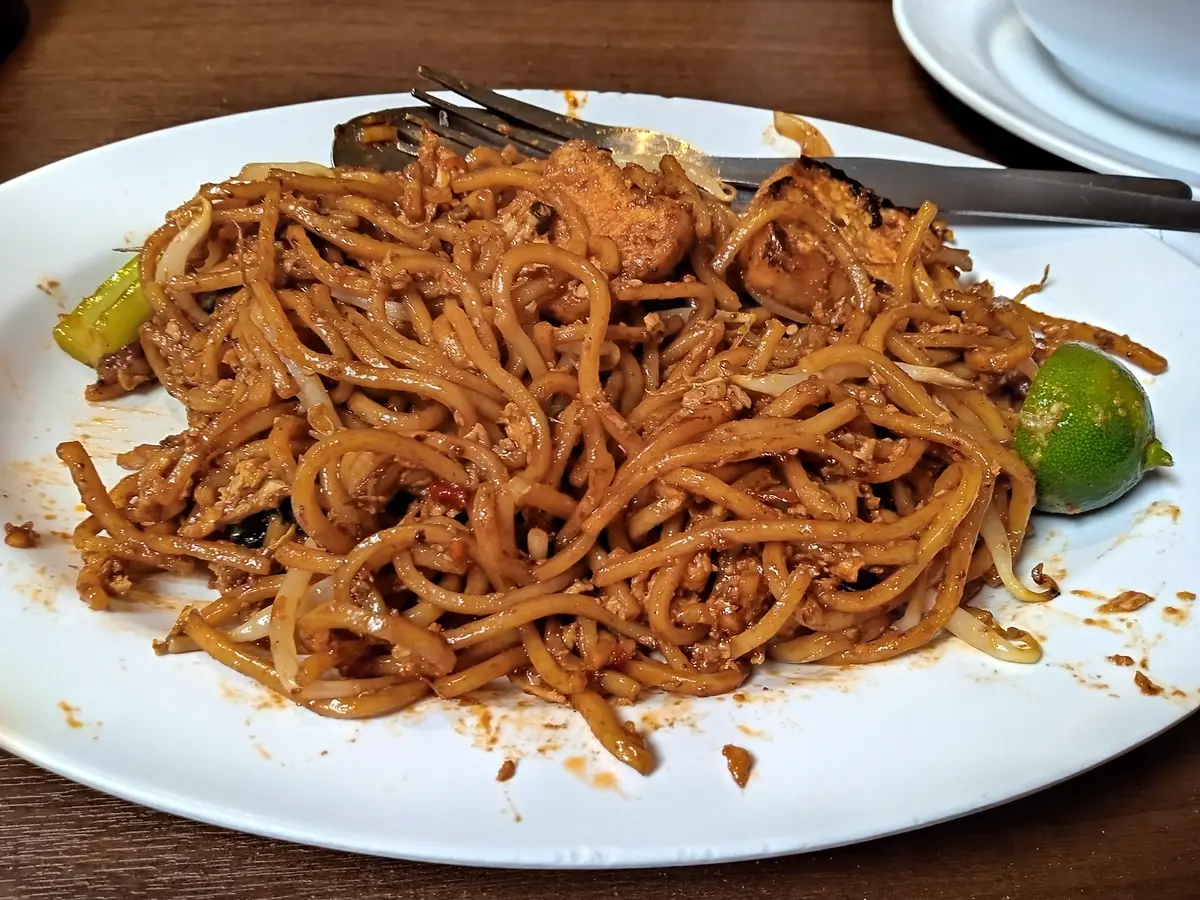 A white plate of mee goreng mamak, stir-fried noodles with pieces of meat, served with a wedge of lime