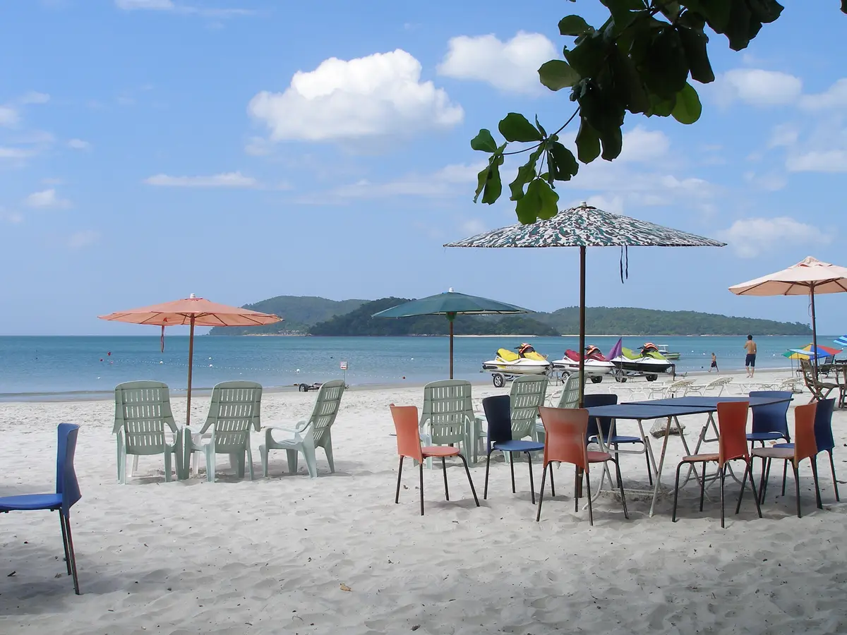 Colorful chairs, a table and beach umbrellas on the white sand at Pantai Cenang in Langkawi, with jet skis on the water and green hills across the bay
