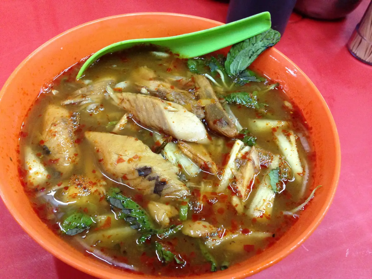 An orange bowl of assam laksa, a sour fish broth with chunks of flaked fish, mint leaves, sliced vegetables and red chili, with a green spoon