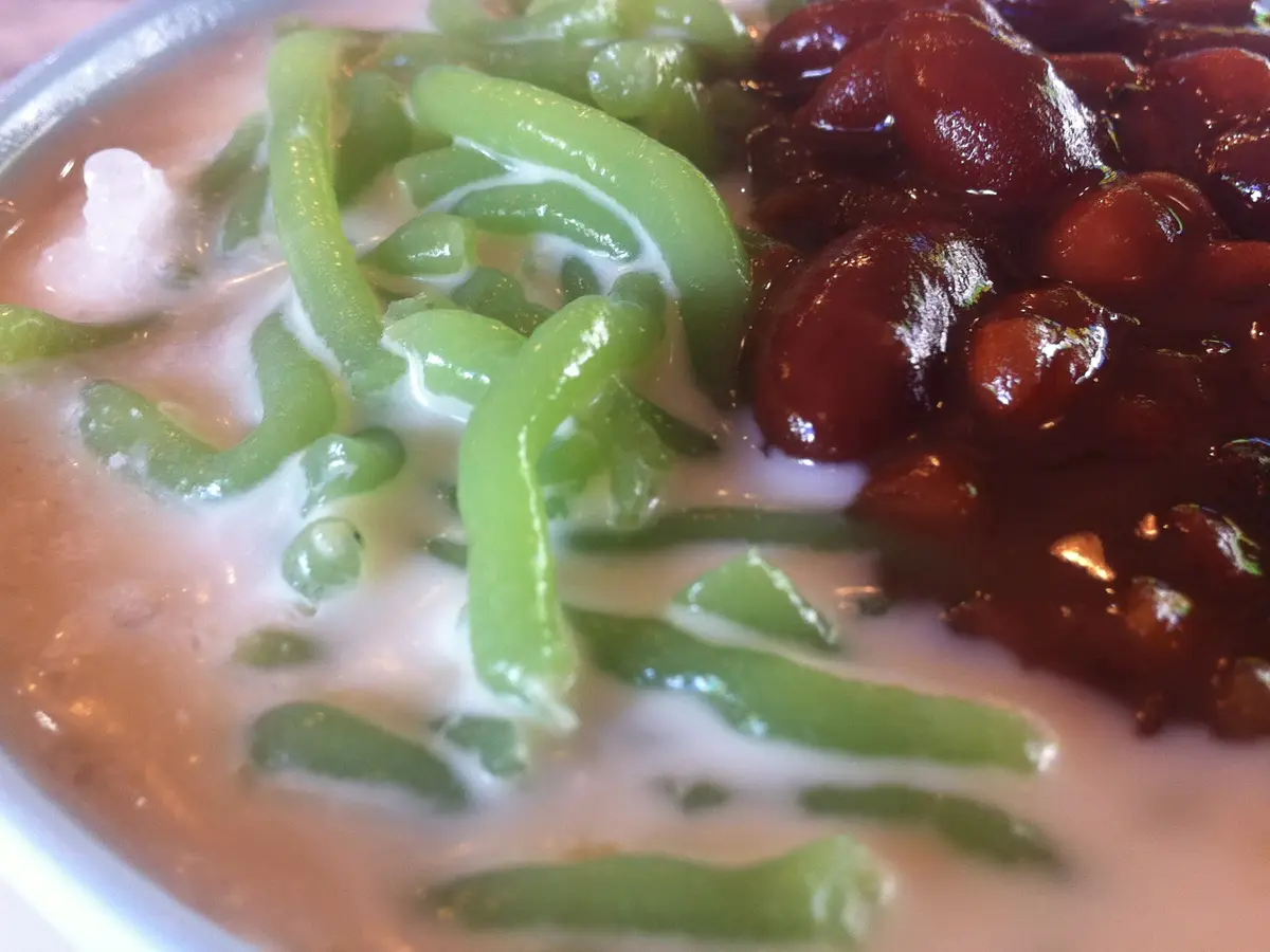 A close view of a bowl of cendol: green jelly strands floating in creamy coconut milk beside a mound of sweet red beans