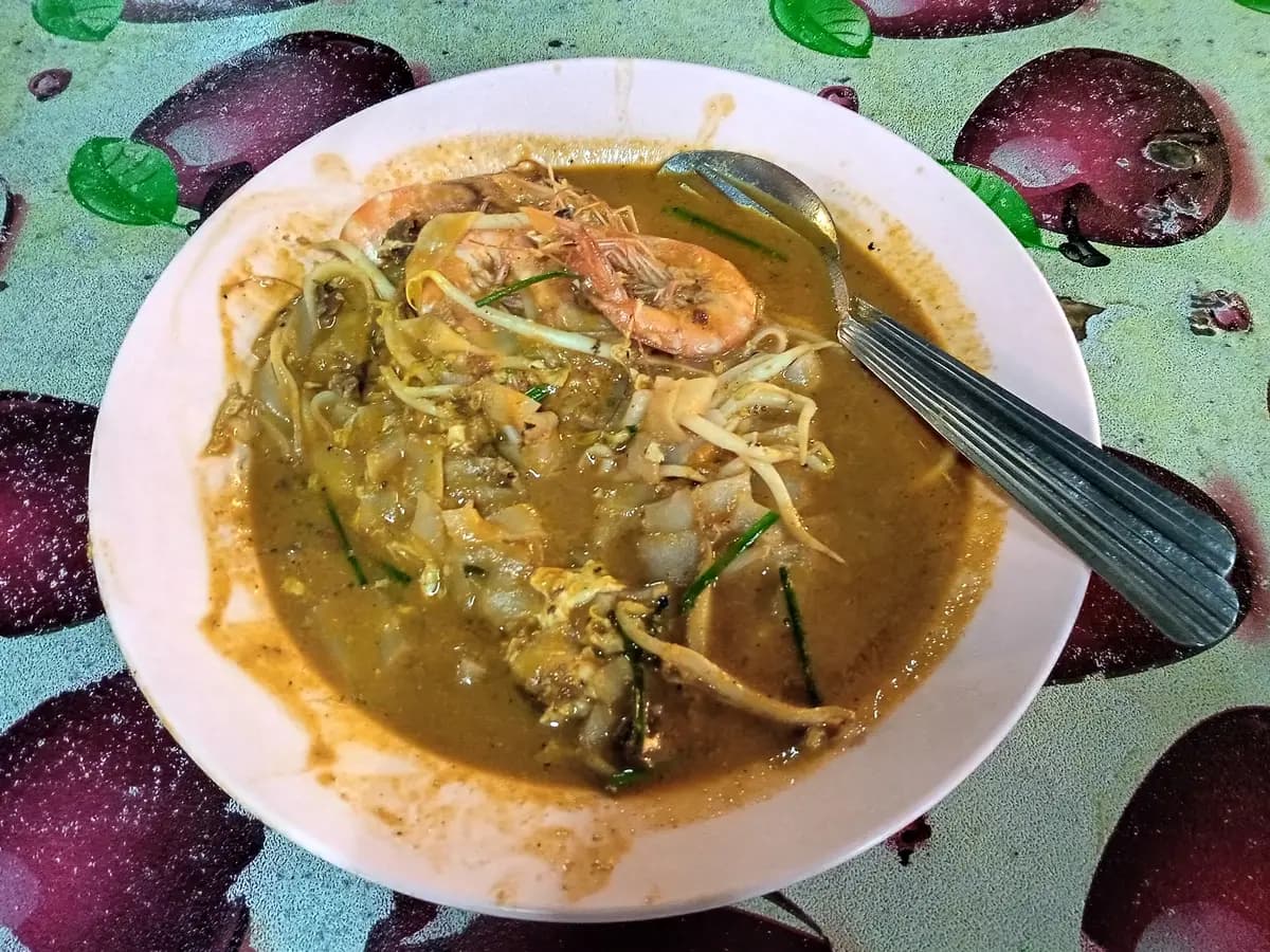 A bowl of char kway teow, flat rice noodles stir-fried with egg, bean sprouts and Chinese chives and topped with a prawn, served with a metal spoon on a patterned tablecloth