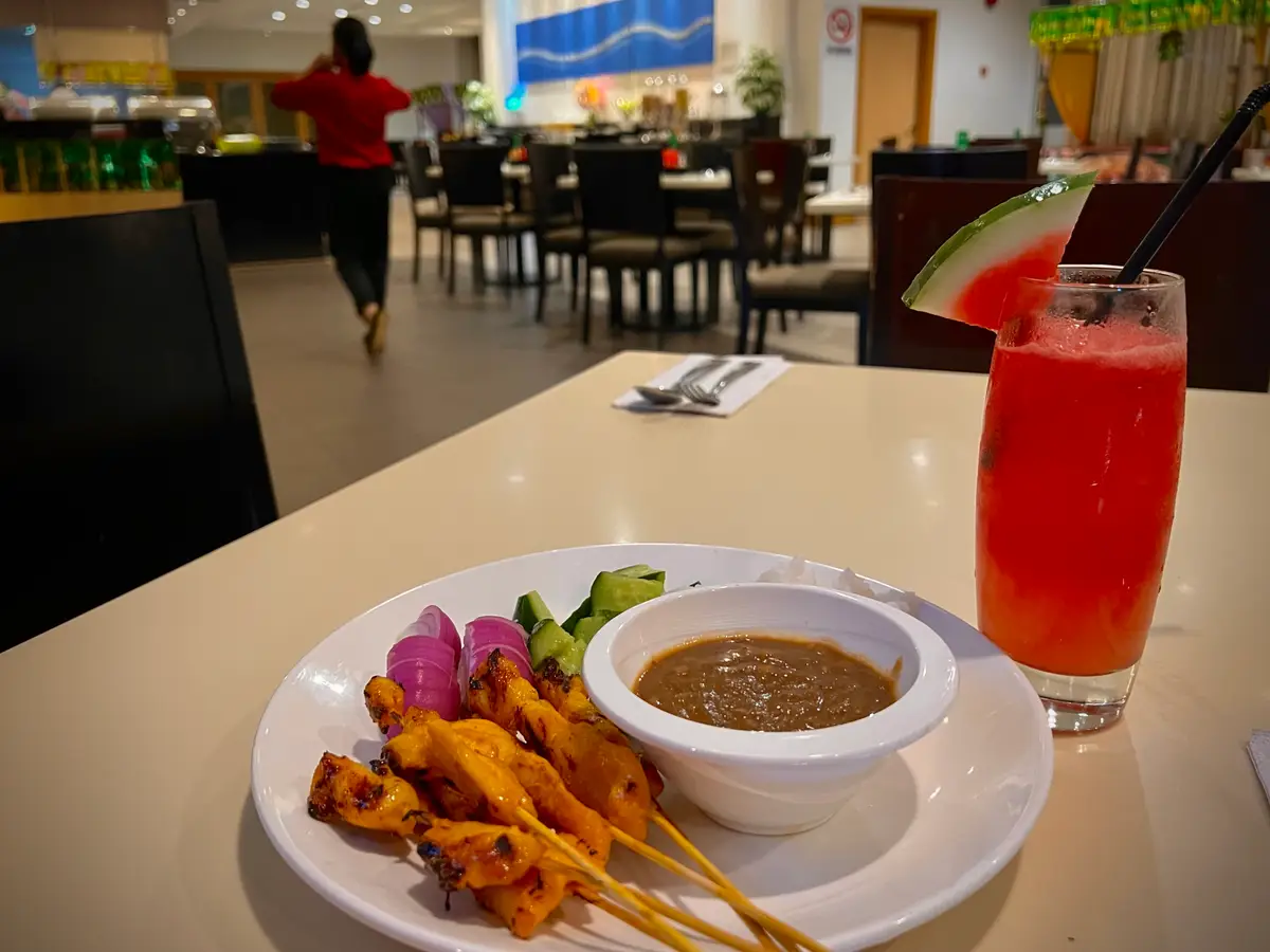 A plate of grilled chicken satay skewers beside a bowl of peanut sauce, with cucumber chunks and red onion, and a glass of watermelon juice in a restaurant