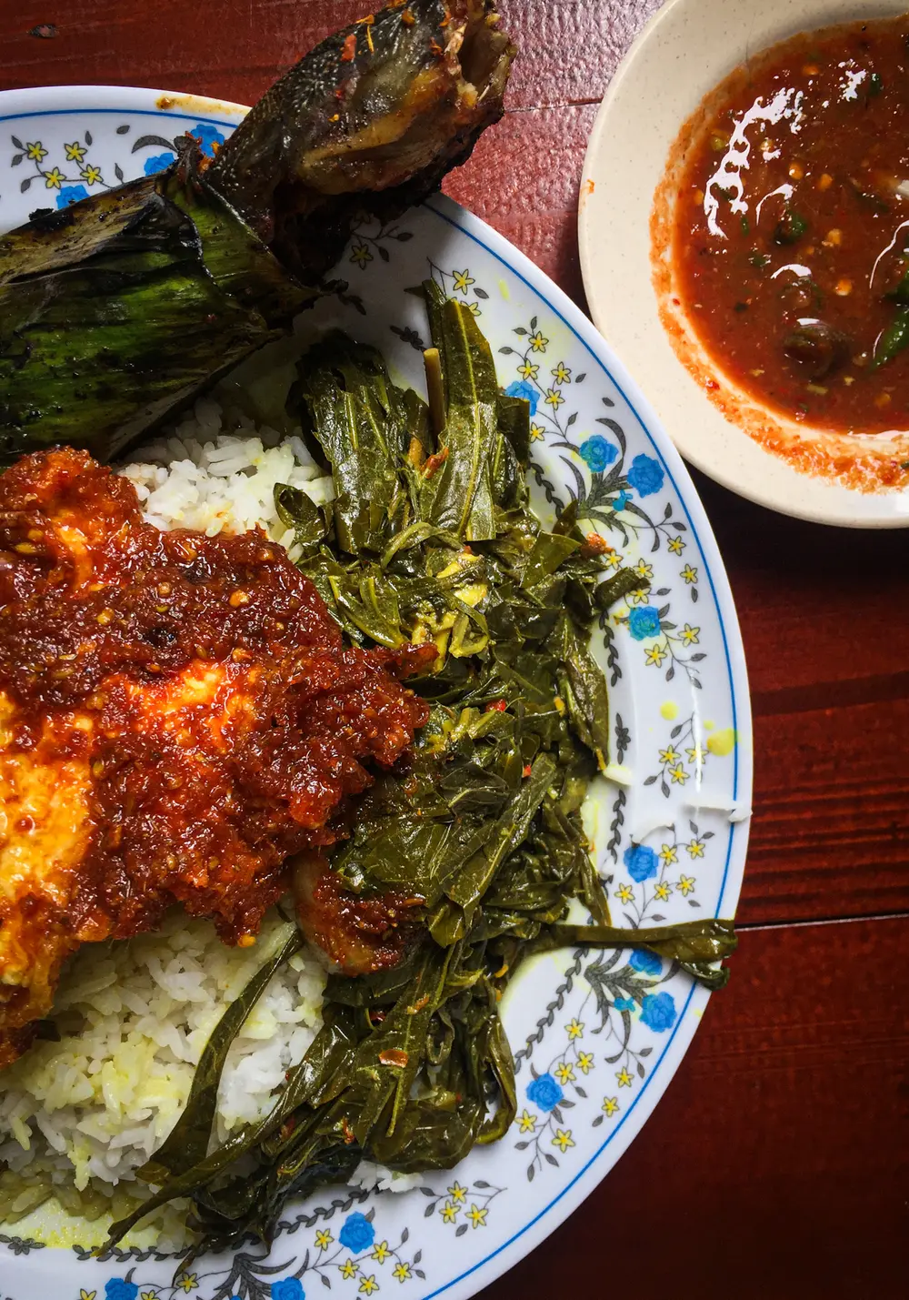 A Malay nasi campur plate seen from above: rice with a glossy red sambal egg, stir-fried greens and a grilled banana-leaf parcel of fish, beside a bowl of sambal