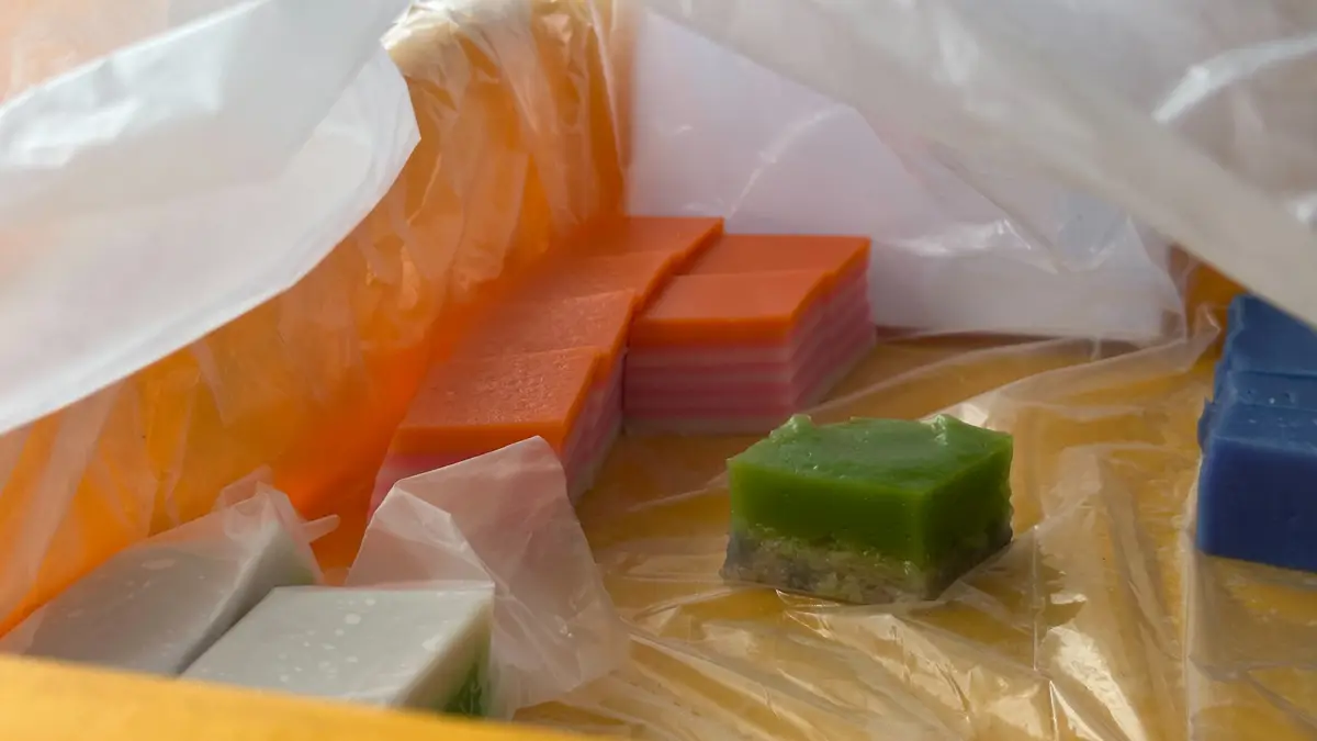 A selection of colorful kuih in plastic bags, including pink and orange layered kuih lapis, a green layered kuih and blue pieces