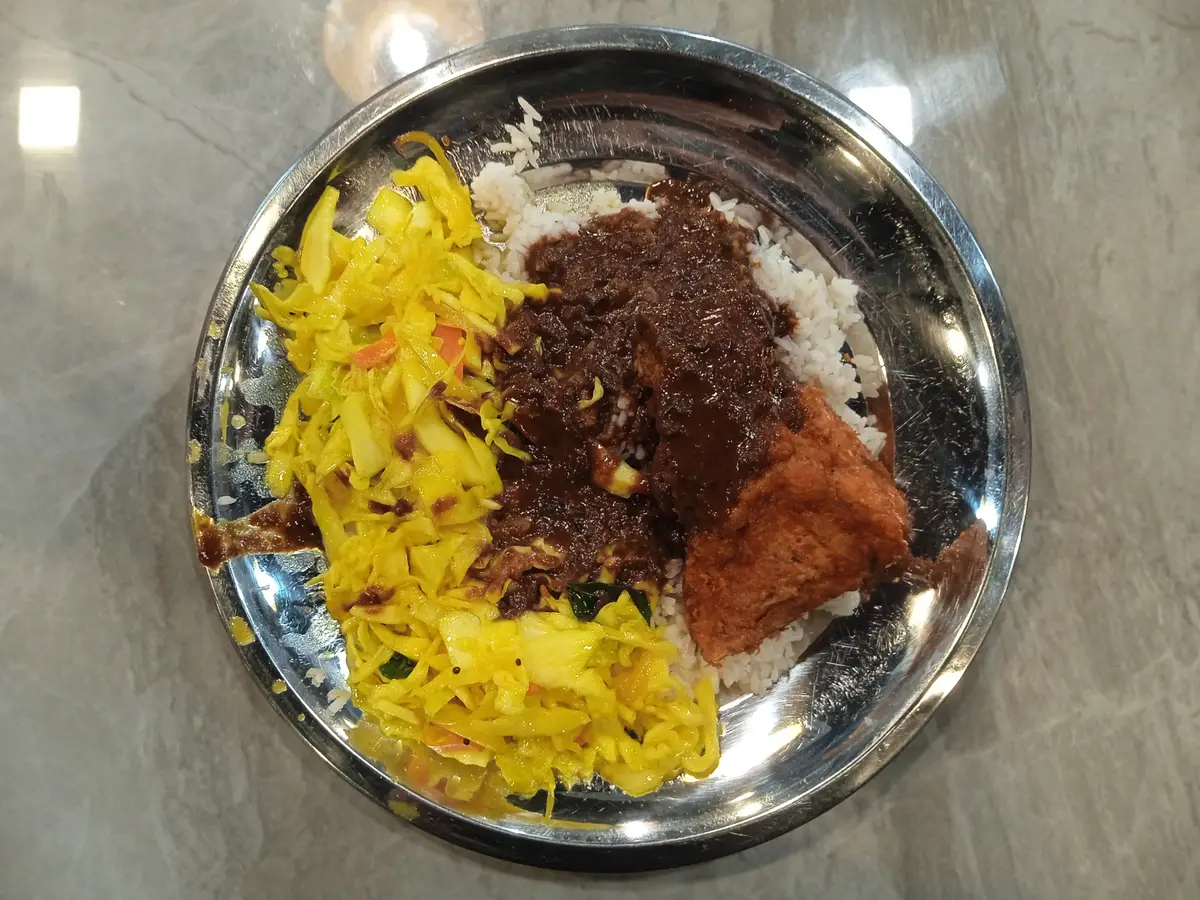 A steel plate of nasi kandar seen from above: white rice with dark curry gravy poured over it, a piece of fried chicken and a side of stir-fried yellow cabbage