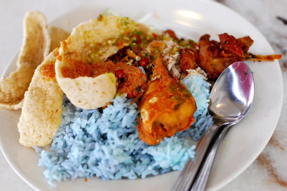 A plate of nasi kerabu with bright blue rice, prawn crackers, a fried main dish, sambal and grated coconut, with a steel spoon on the right