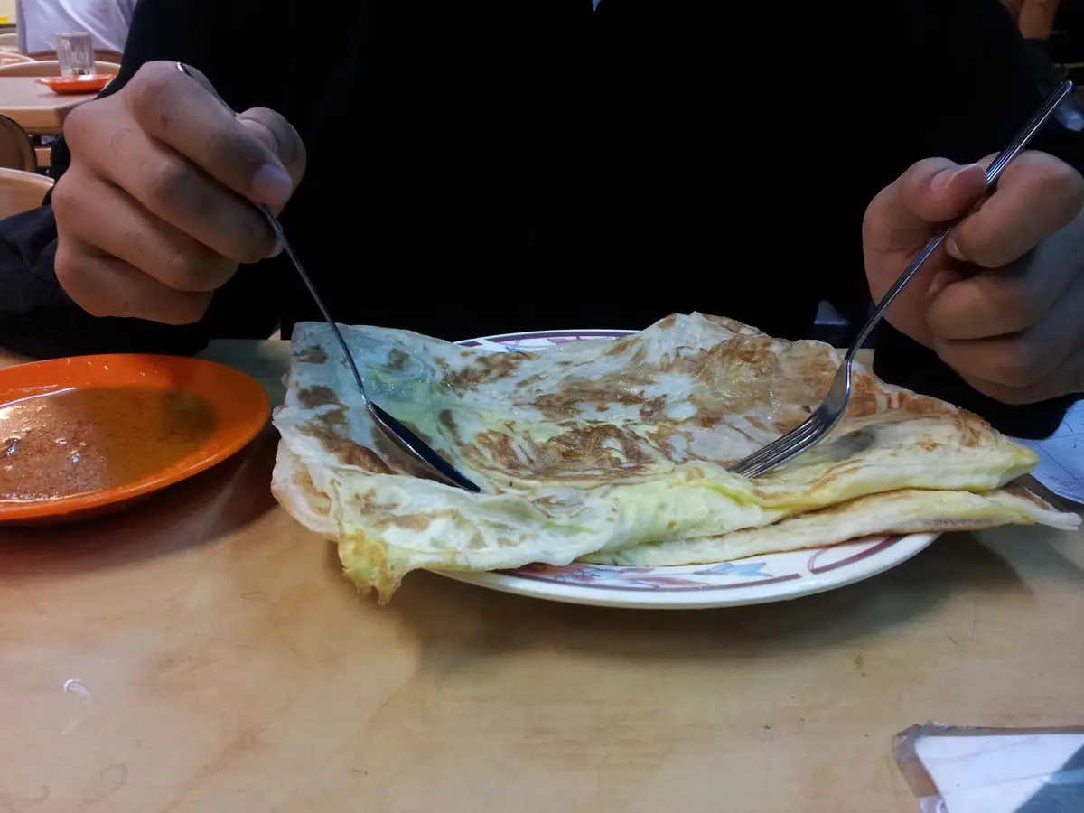 A folded egg roti canai on a patterned plate, with a diner’s hands holding a fork and spoon over it and an orange bowl of curry sauce beside the plate