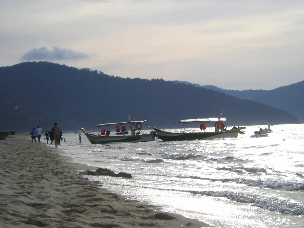 Two small tour boats moored in shallow water off a sandy beach at dusk, with a forested hill behind and people walking along the shore