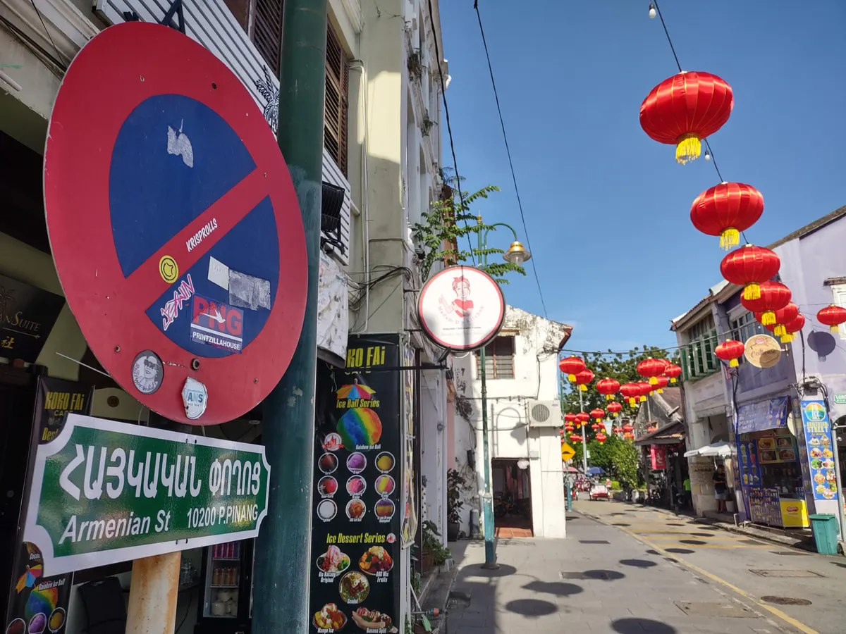Strings of red lanterns hang above Armenian Street in George Town between old shophouses, beside a green street sign written in Armenian and English