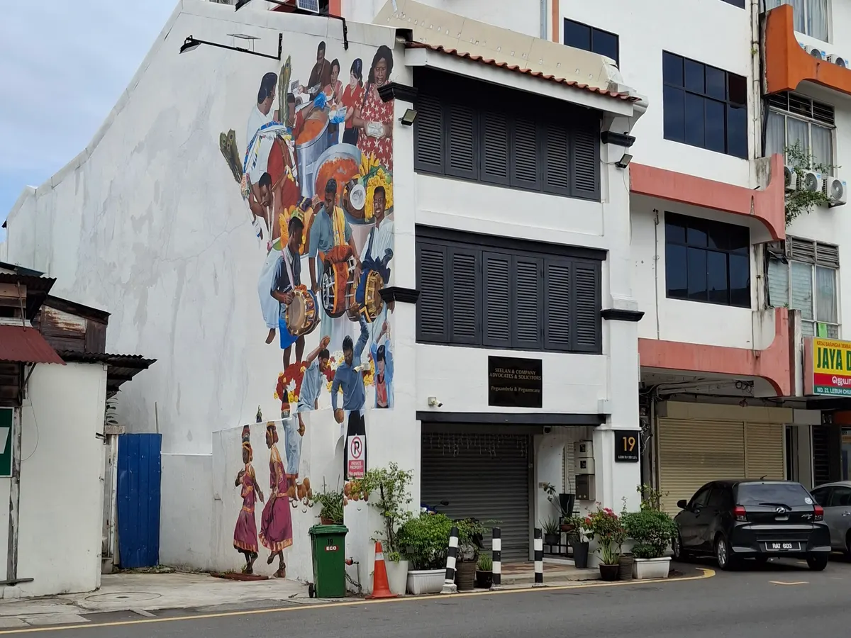A white building on a George Town street with a colorful mural of dancers and musicians painted on its side wall, beside a road with parked cars