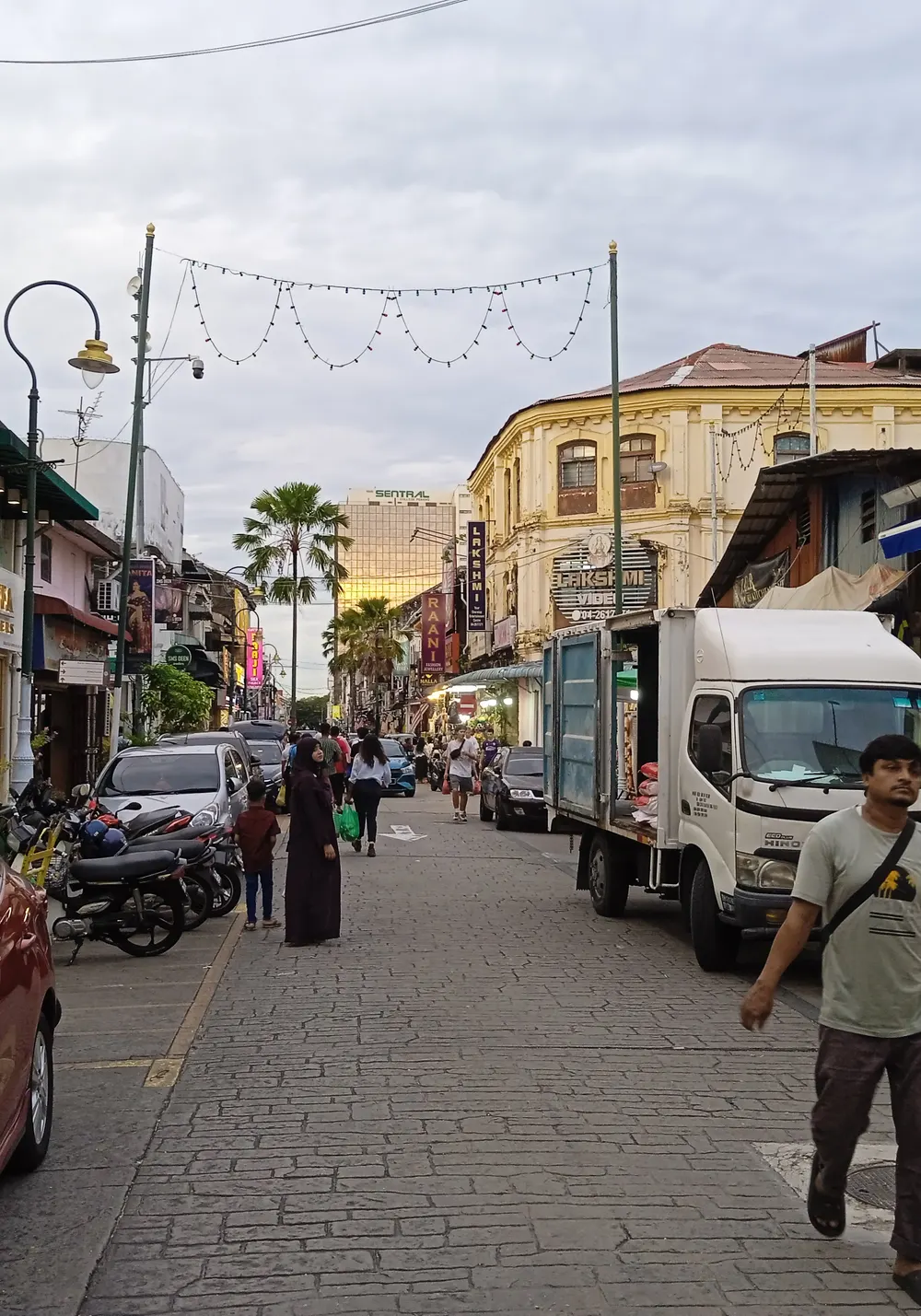 A busy pedestrian street in George Town, Penang, with heritage shophouses, shop signs, strings of lights and people walking under an evening sky