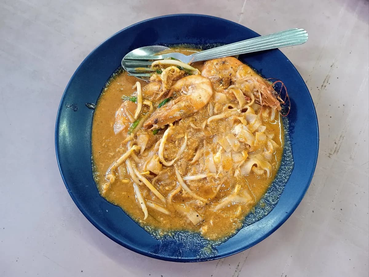 A blue plate of Penang char kway teow, flat rice noodles with prawns, bean sprouts and green chives in an orange sauce, with a fork and spoon