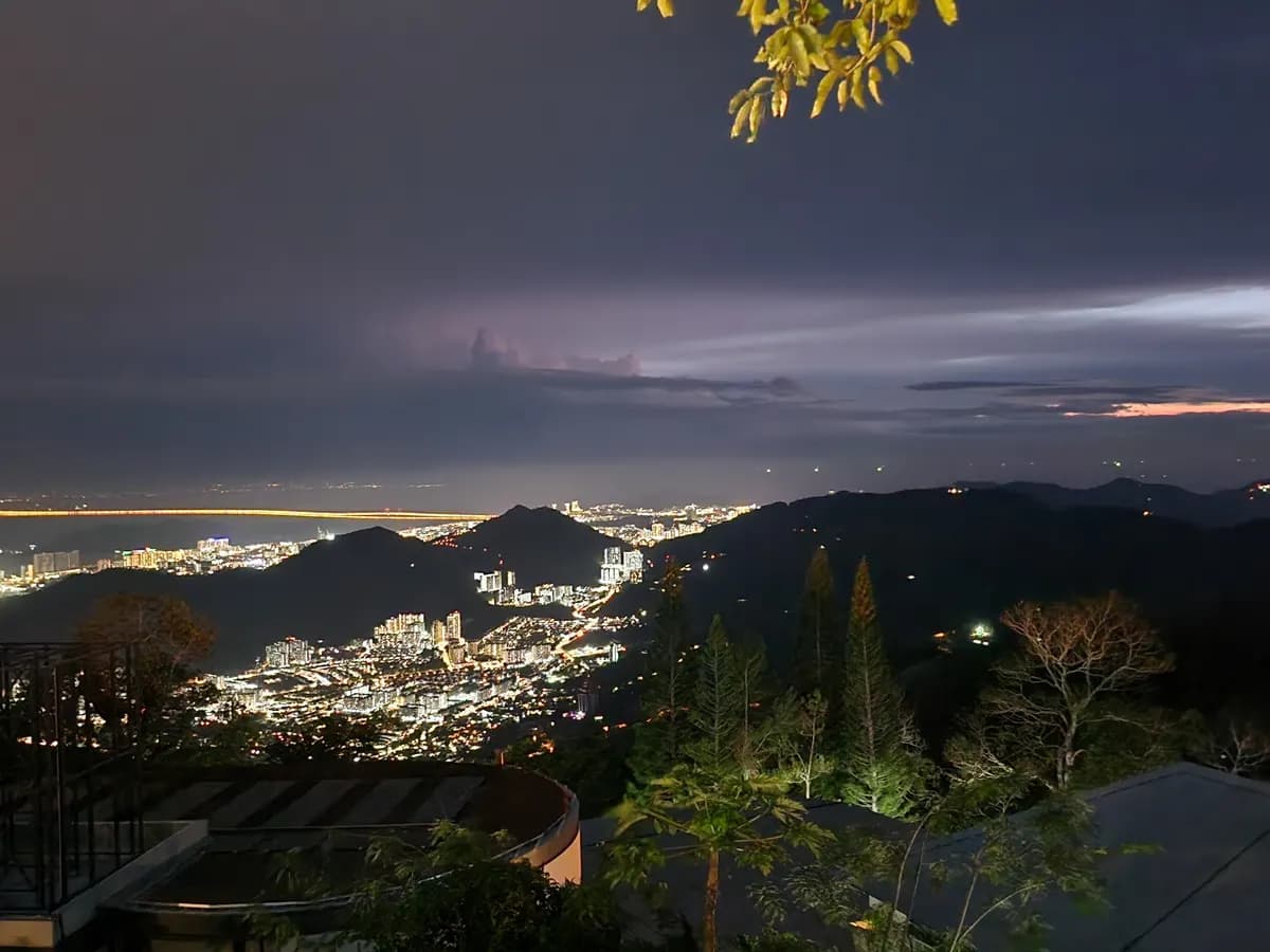 Evening view from Penang Hill over dark forested ridges and the glowing lights of George Town, with clouds and a pink glow above the water