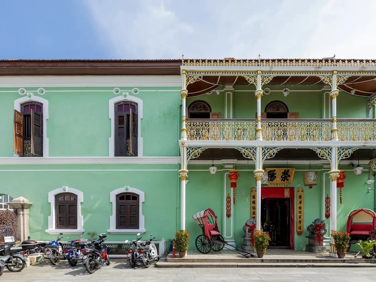 The pale green facade of Pinang Peranakan Mansion with a gilded iron balcony, shuttered windows, red lanterns and a decorative rickshaw at the entrance