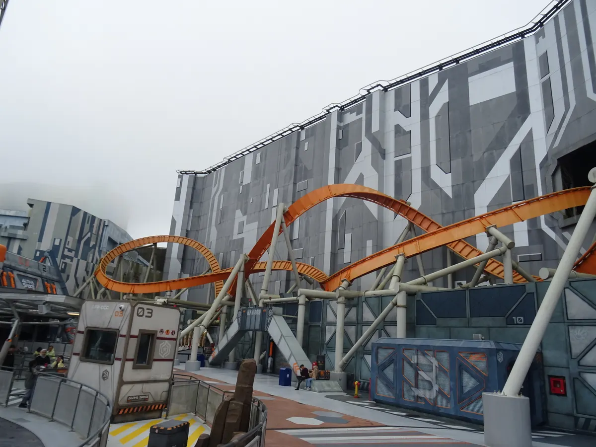 An orange looping roller coaster curving around a grey, futuristic building in the Andromeda Base area of Genting SkyWorlds, under an overcast mountain sky