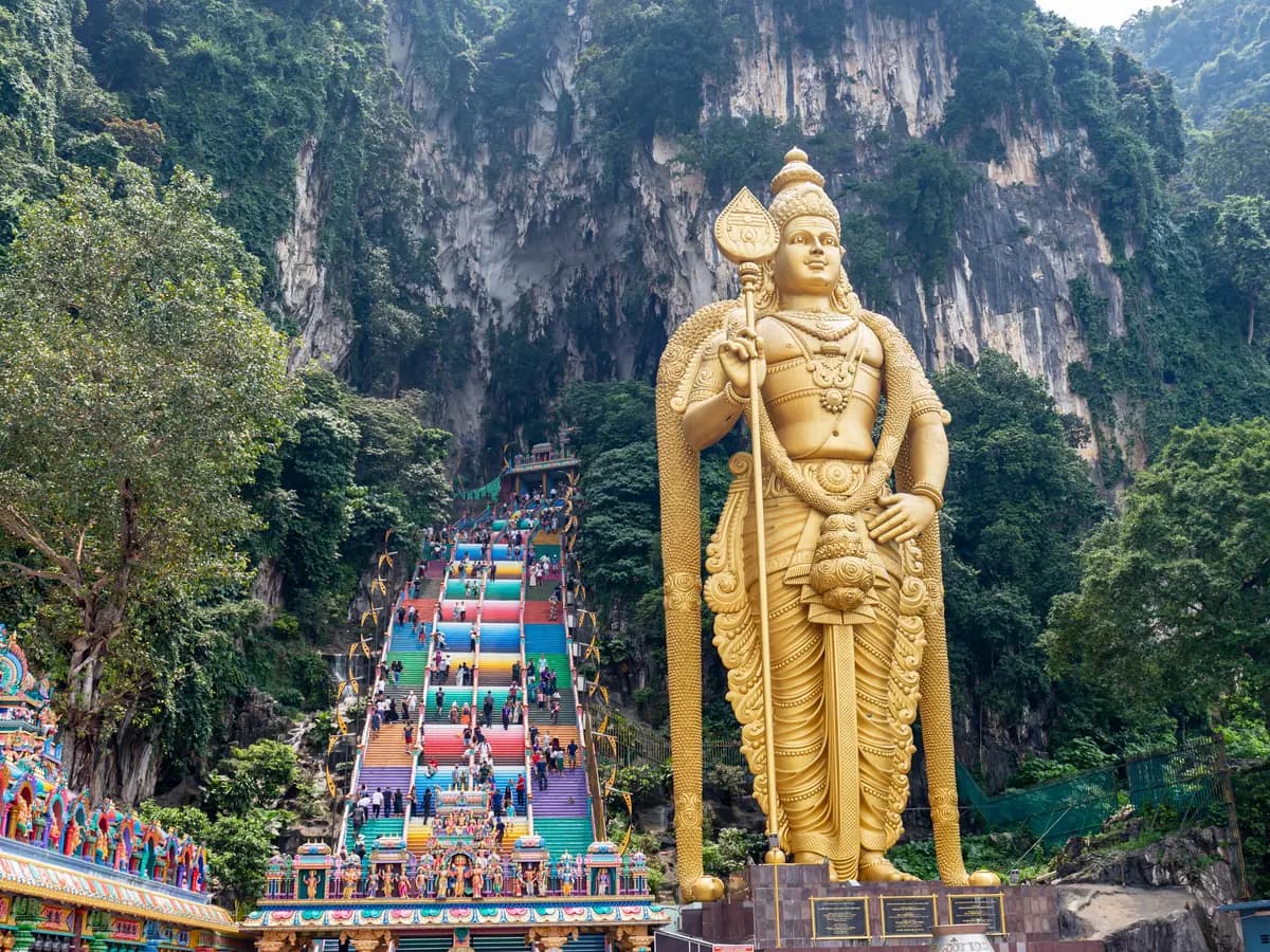 The golden Murugan statue beside the colourful stairway leading up to the Batu Caves temple, below a limestone cliff covered in green forest