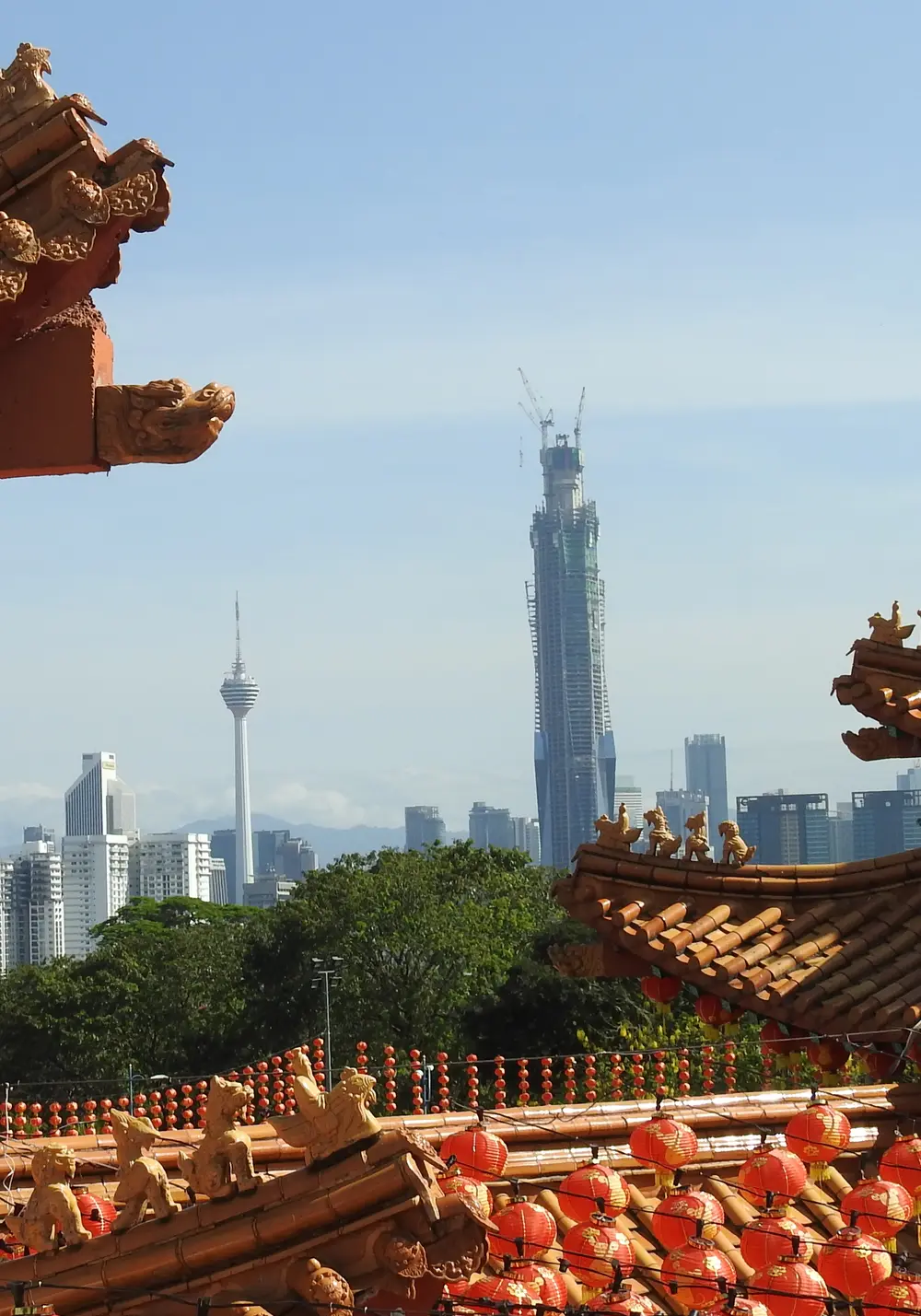 Orange-tiled temple roofs hung with red lanterns at Thean Hou Temple in Kuala Lumpur, with the KL Tower and a tall tower under construction rising beyond the city’s trees and apartment blocks